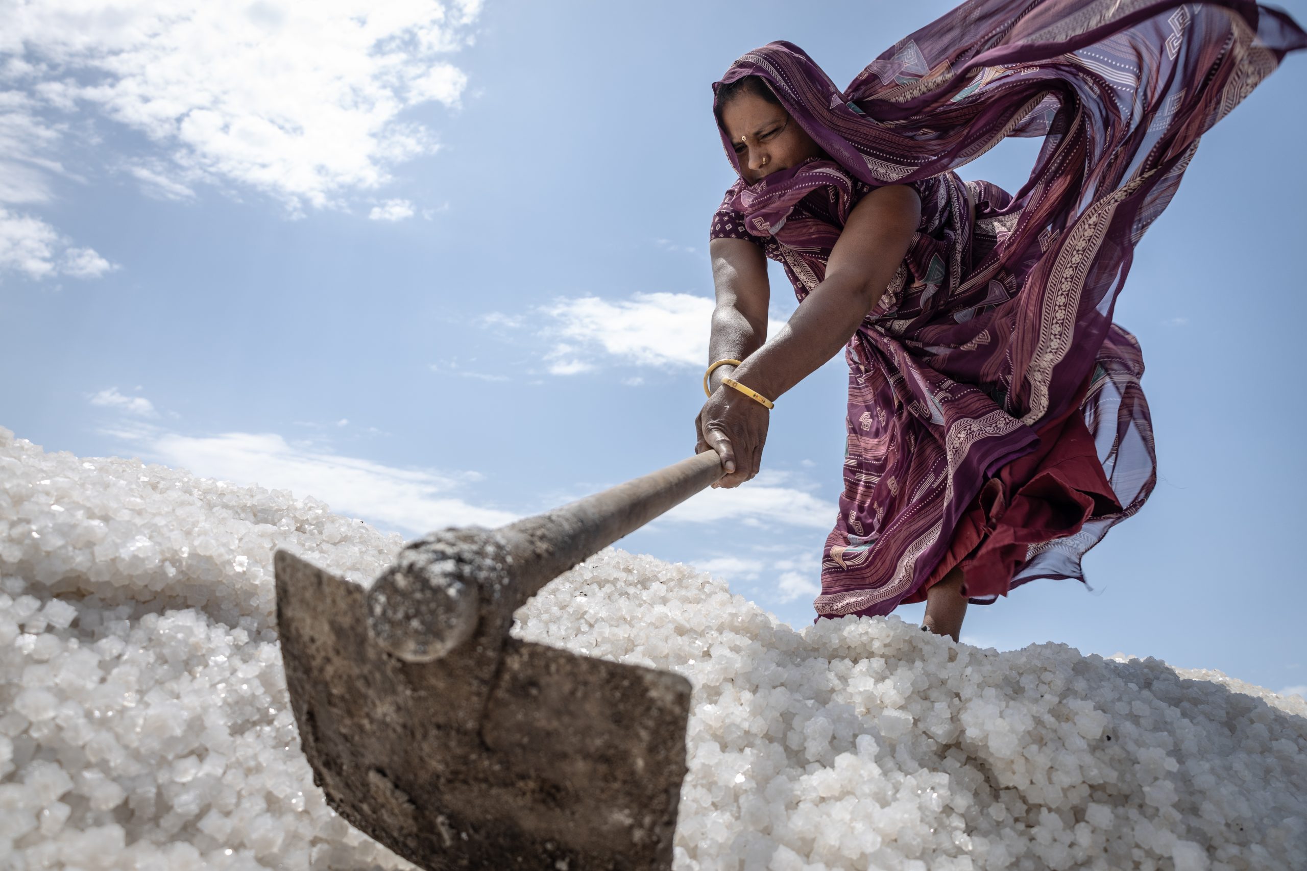 Low angle view of a worker drying salt, moving the salt with a spade. It is a hot day with strong sunlight and their clothing is blowing in the wind.